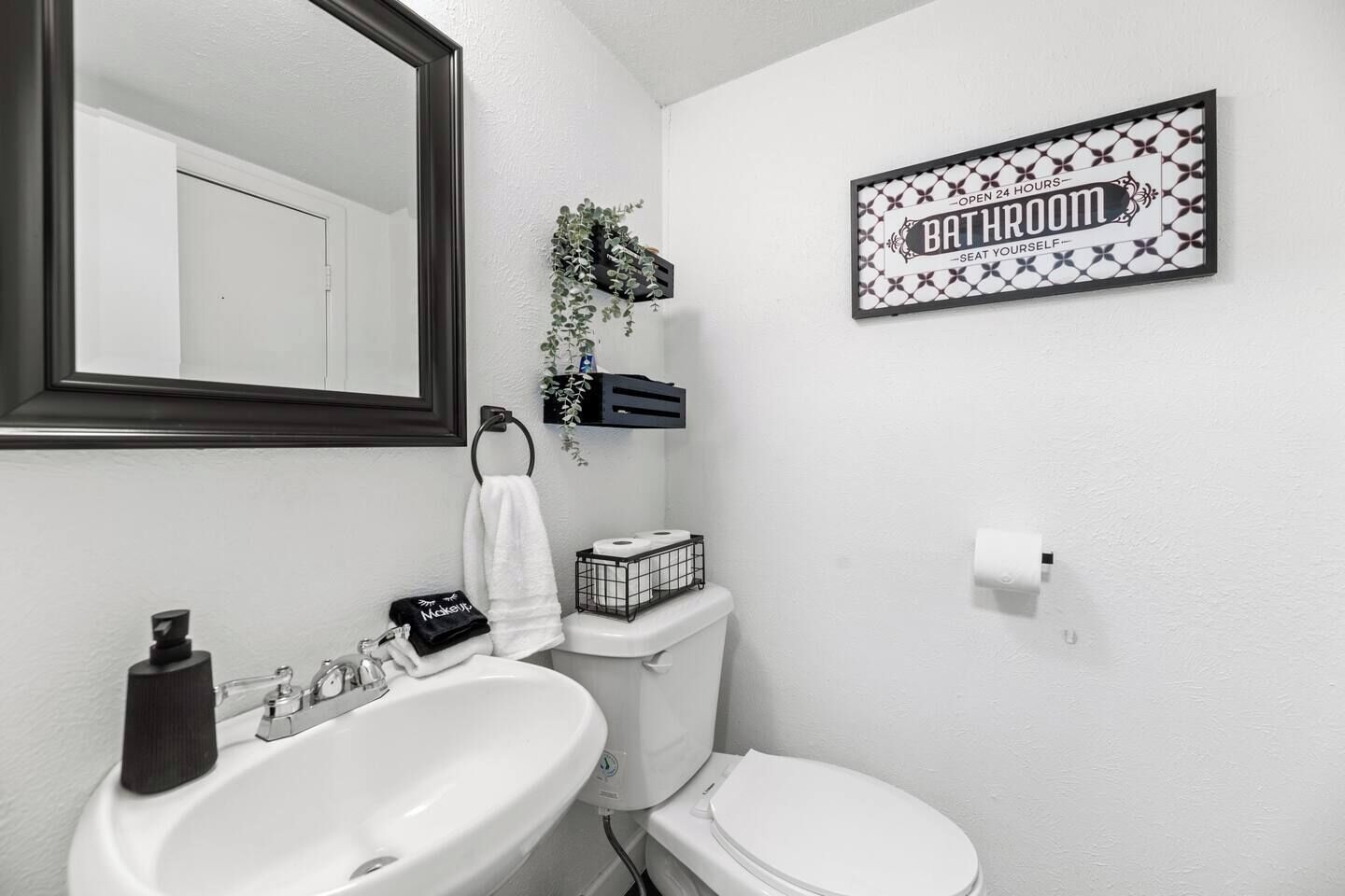 Un baño con un lavamanos blanco, un inodoro, un espejo y un letrero que dice 'BATHROOM - OPEN 24 HOURS - SEAT YOURSELF'.