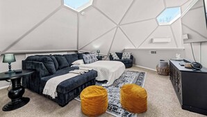 A modern, geometric bedroom with a high ceiling and skylights, a dark sofa, a bed with a white duvet, and a small desk area. - Dome Manor / Pool + Hot Tub + Two Game Rooms (Bedford)