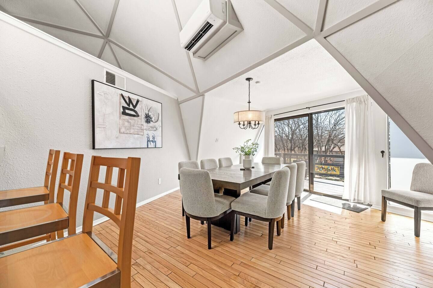 A dining area with a wooden table and chairs, a chandelier, and a large window with a view of trees.