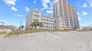 Ein Strandhotel mit mehreren Balkonen, ein Sandstrand im Vordergrund und ein hohes Gebäude im Hintergrund.
