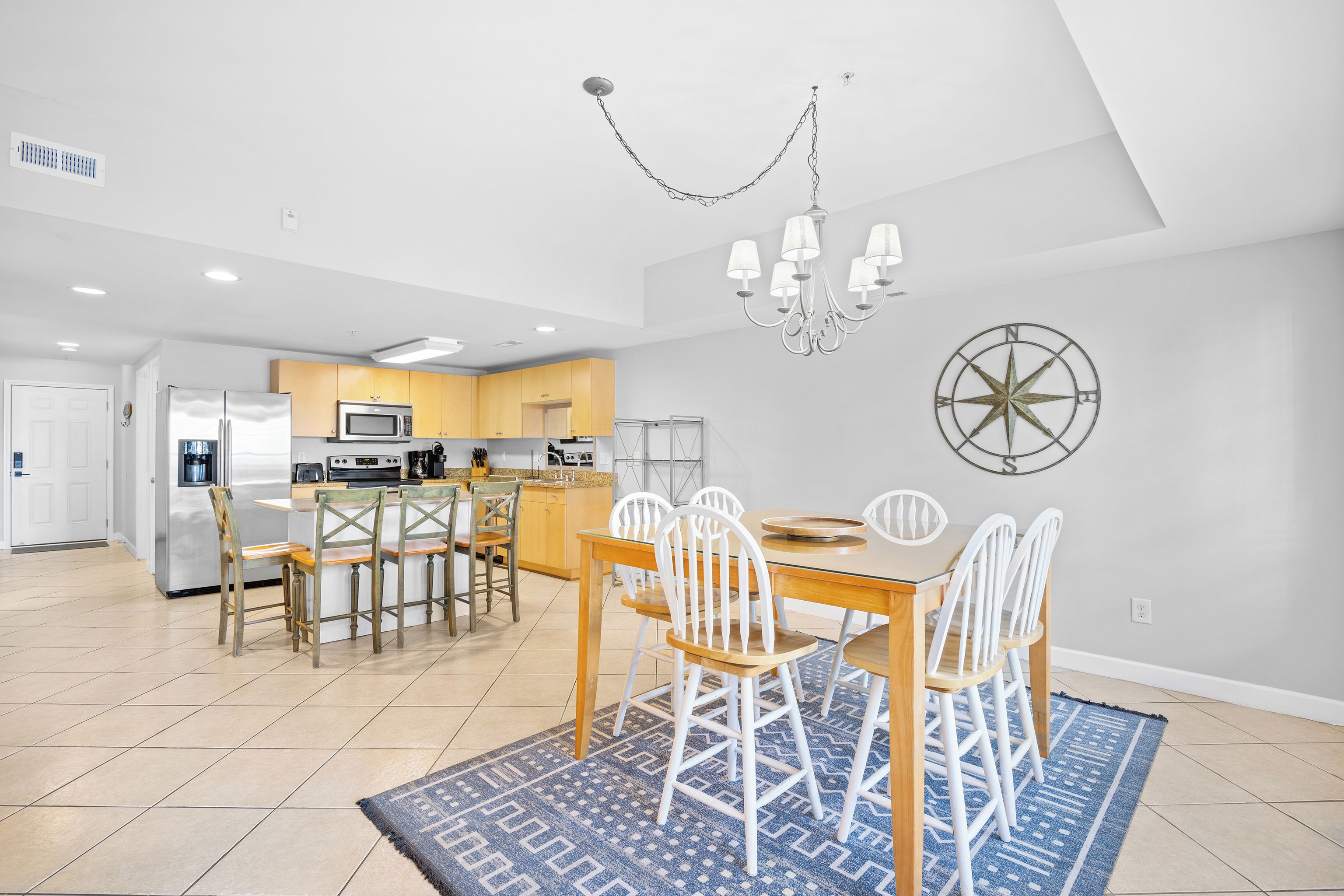 A modern kitchen with a dining area, featuring a chandelier, a compass wall decoration, and a blue patterned rug.