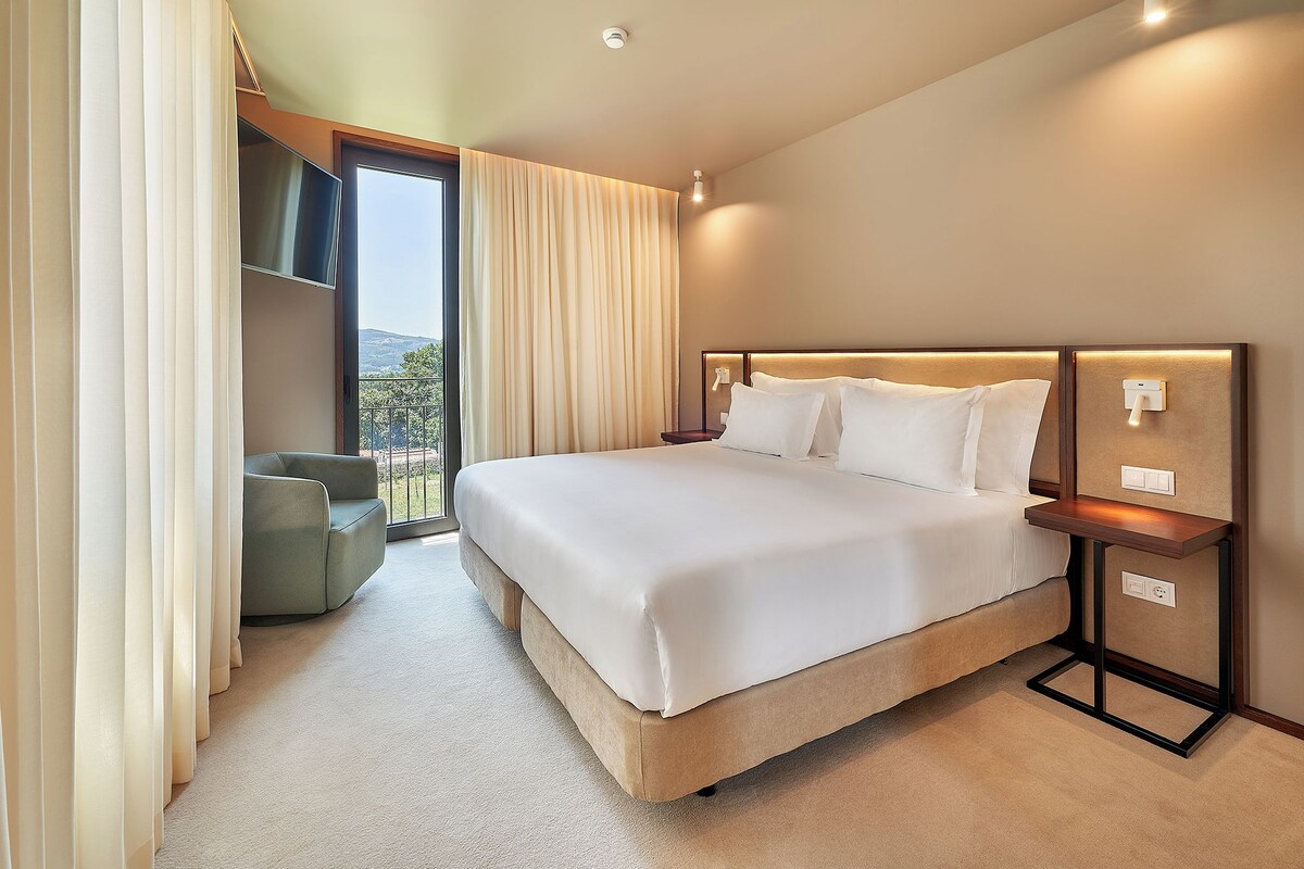A hotel room with a large bed, a nightstand, a chair, and a view of the outdoors.