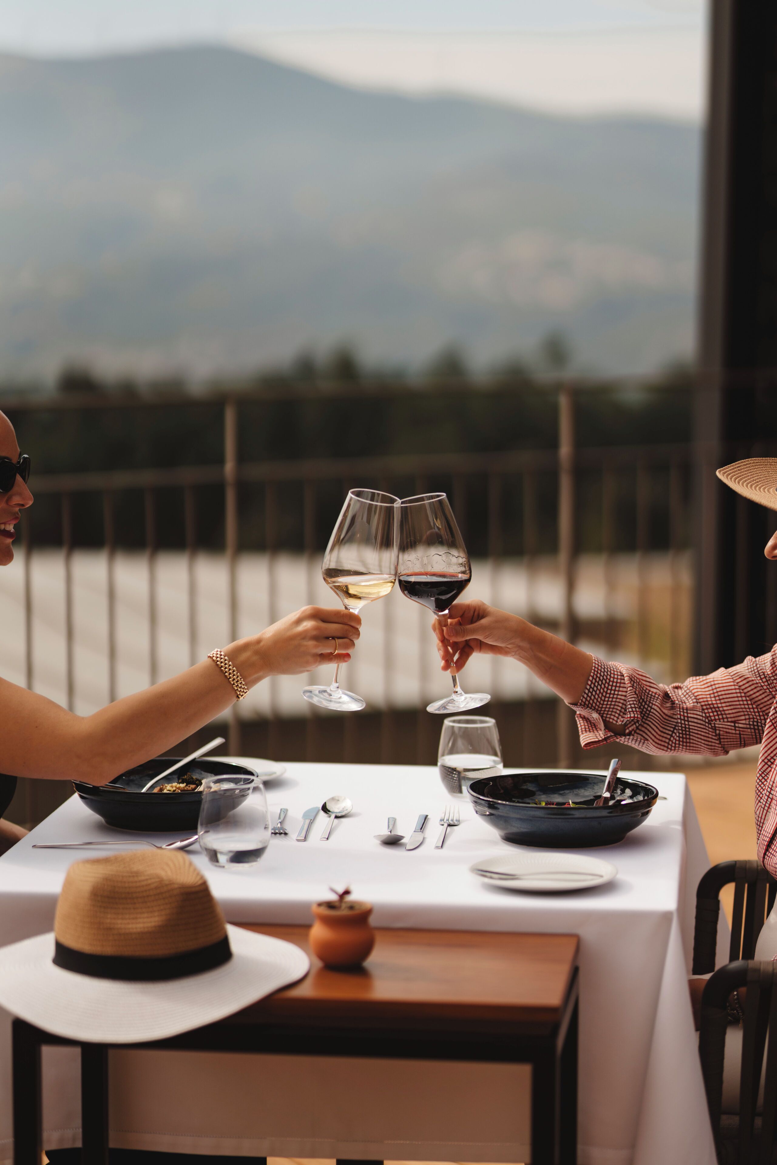 A dining table with a white tablecloth, two wine glasses, a hat, and a bowl of food.