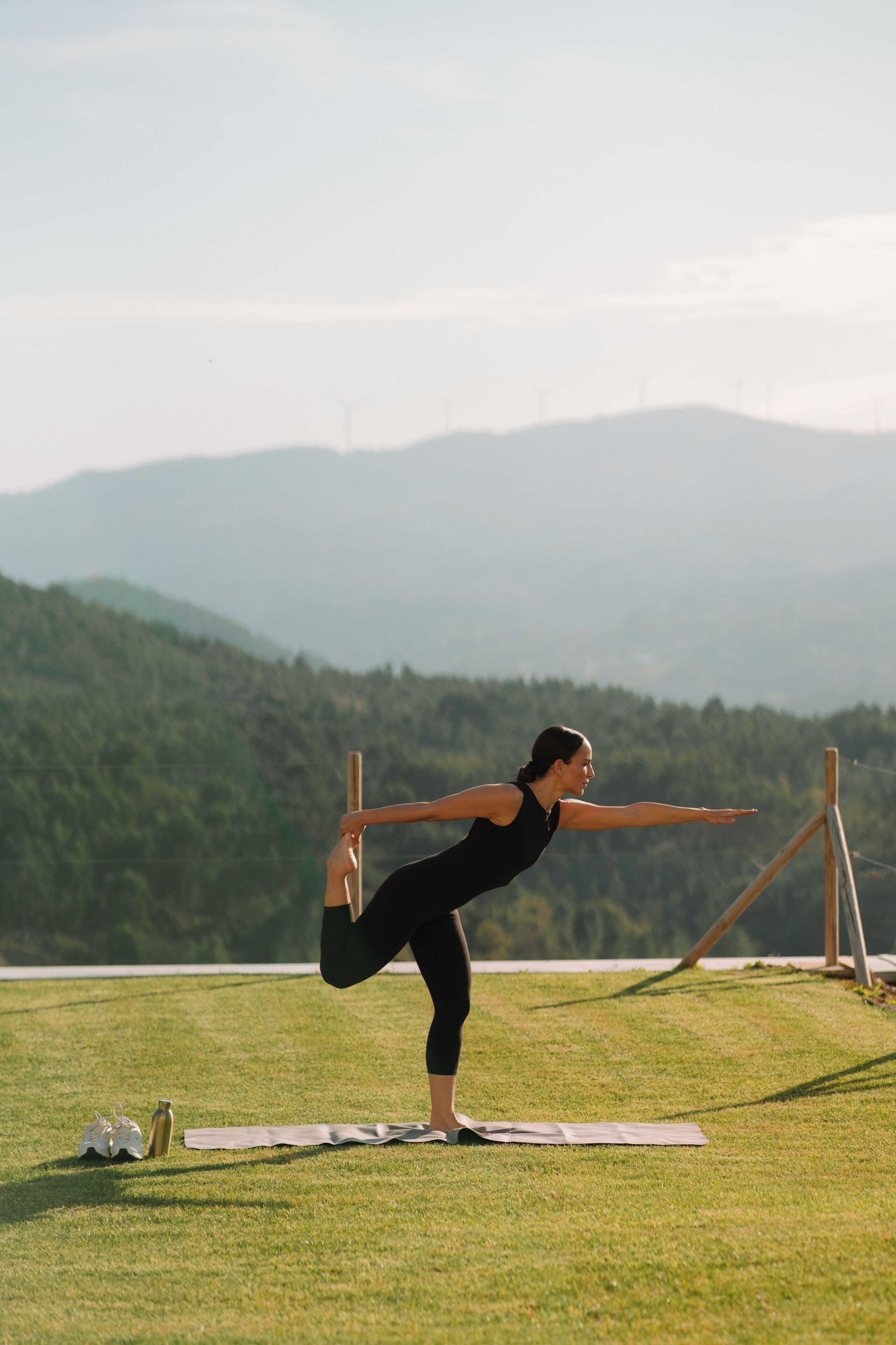 Uma pessoa praticando uma postura de yoga em um tapete ao ar livre, com montanhas ao fundo.