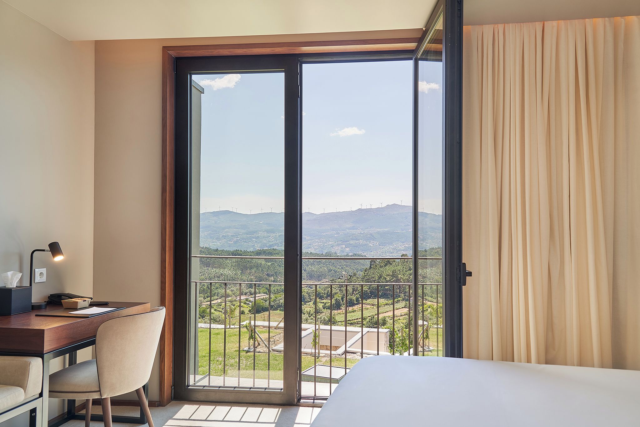 Ein Hotelzimmer mit einem Bett, einem Schreibtisch und einem Stuhl. Ein großes Fenster bietet Blick auf eine gebirgige Landschaft und Windkraftanlagen.