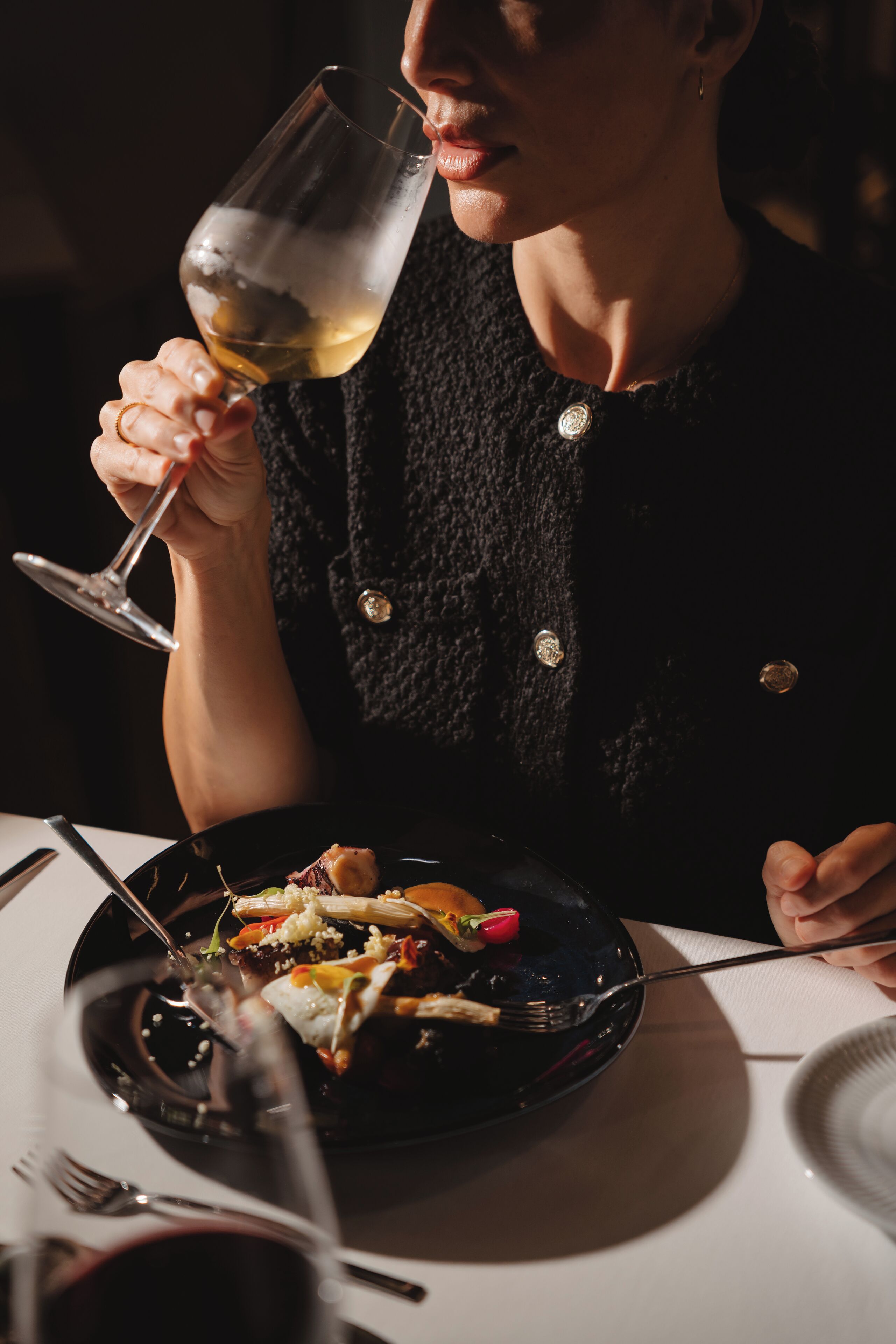 A person holding a wine glass at a dining table with a plate of food.