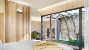 Une chambre moderne avec un grand lit, un fauteuil vert et une vue sur un mur de pierre et la verdure environnante.