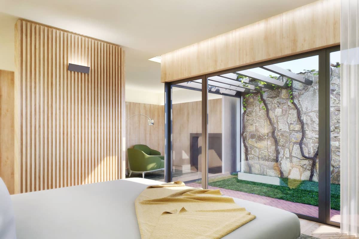 A modern bedroom with a large bed, a green chair, and a view of a stone wall and greenery outside.