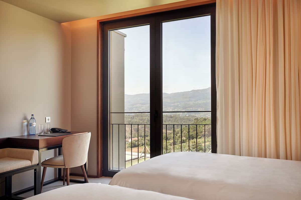 A hotel room with a bed, a desk with a chair, a telephone, and a window with curtains.