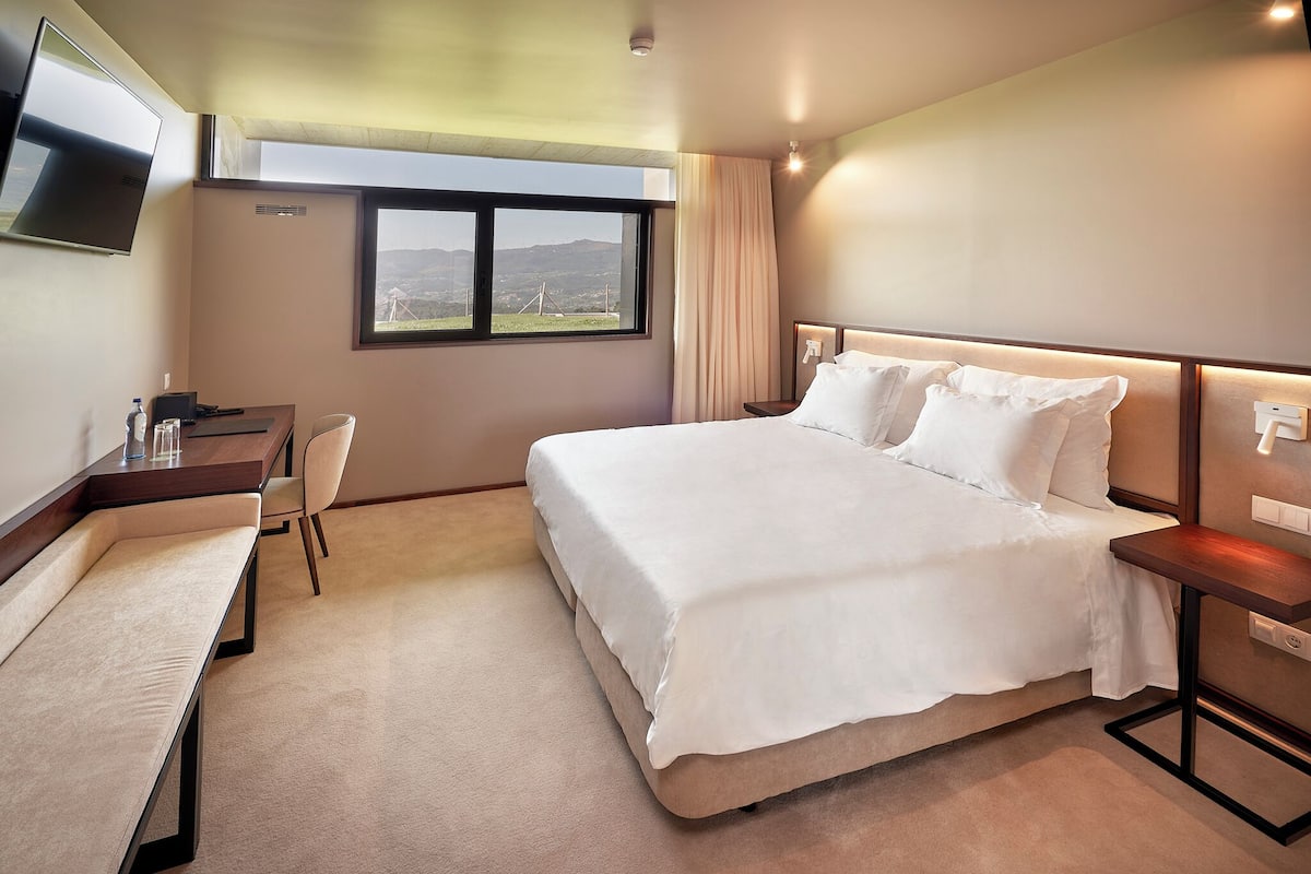 A hotel room with a large bed, a desk with a chair, a flat-screen TV mounted on the wall, and a window with a view of the outdoors.