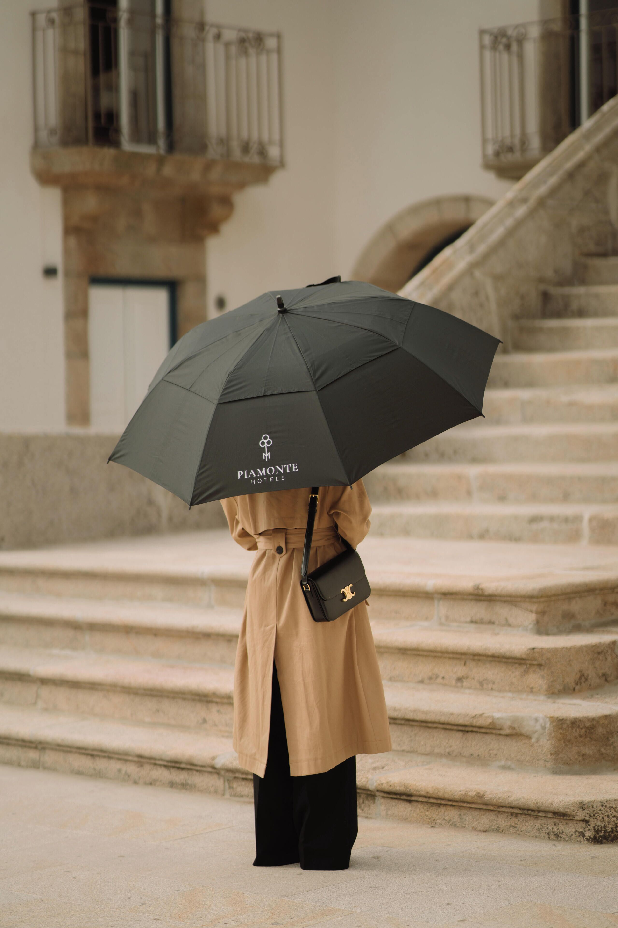A person holding a black umbrella with a logo, standing on stone steps in front of a building with a balcony.