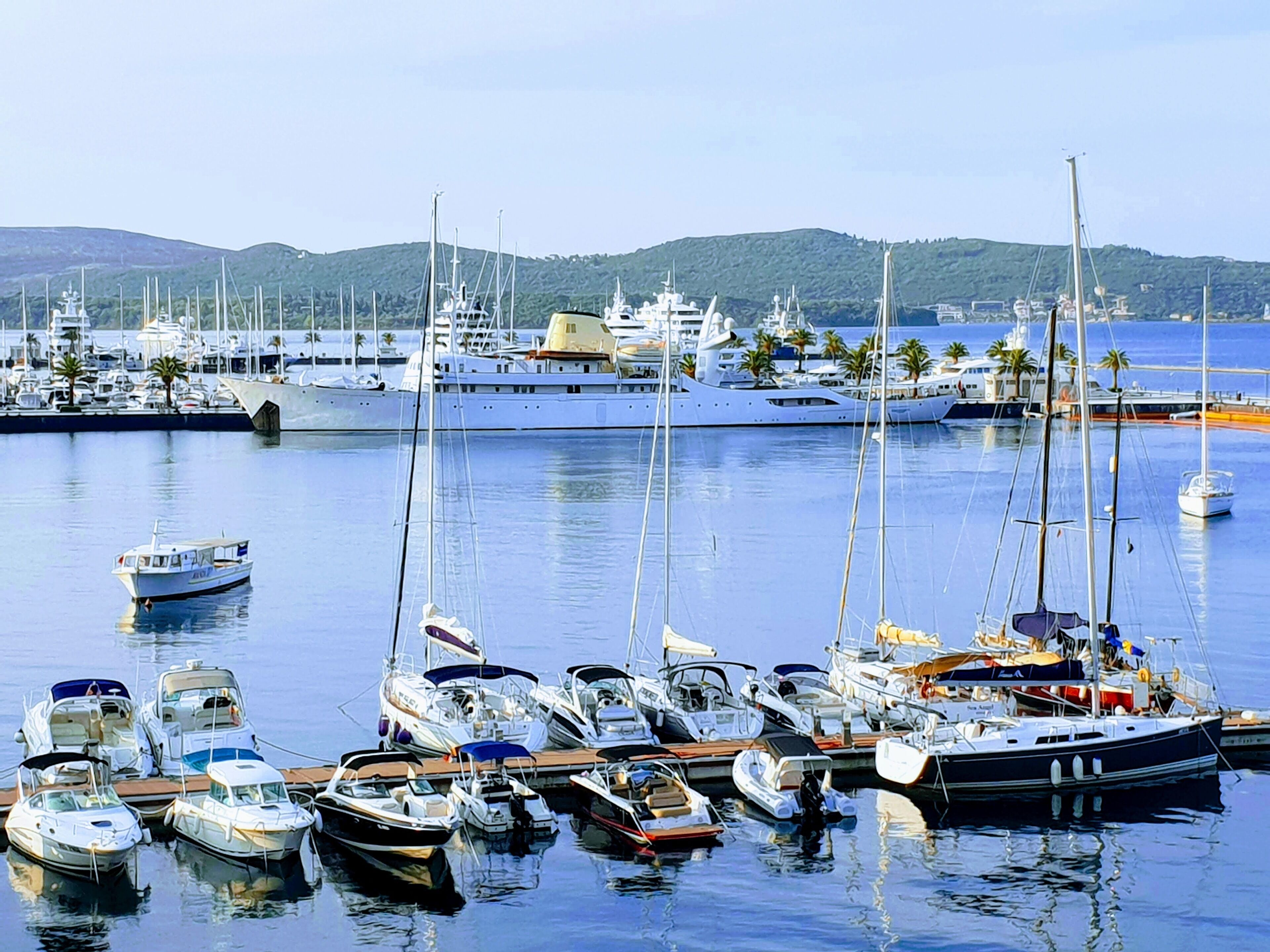 Un port de plaisance où sont amarrés différents types de bateaux, notamment des voiliers et des bateaux à moteur, avec en arrière-plan des collines et un ciel dégagé.