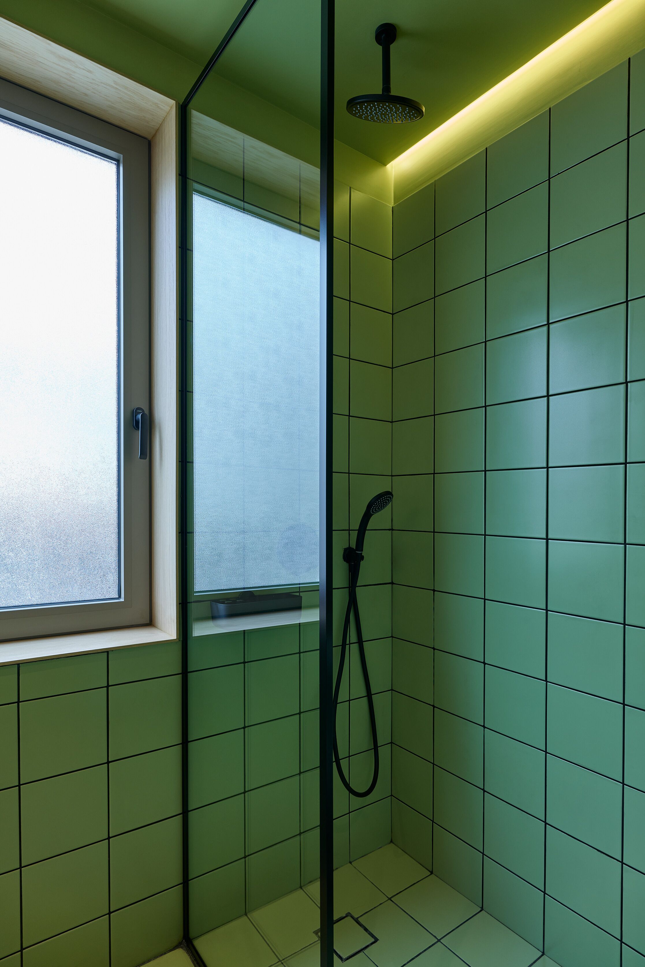 A small, modern shower room with green tiles, a window, and a handheld showerhead.