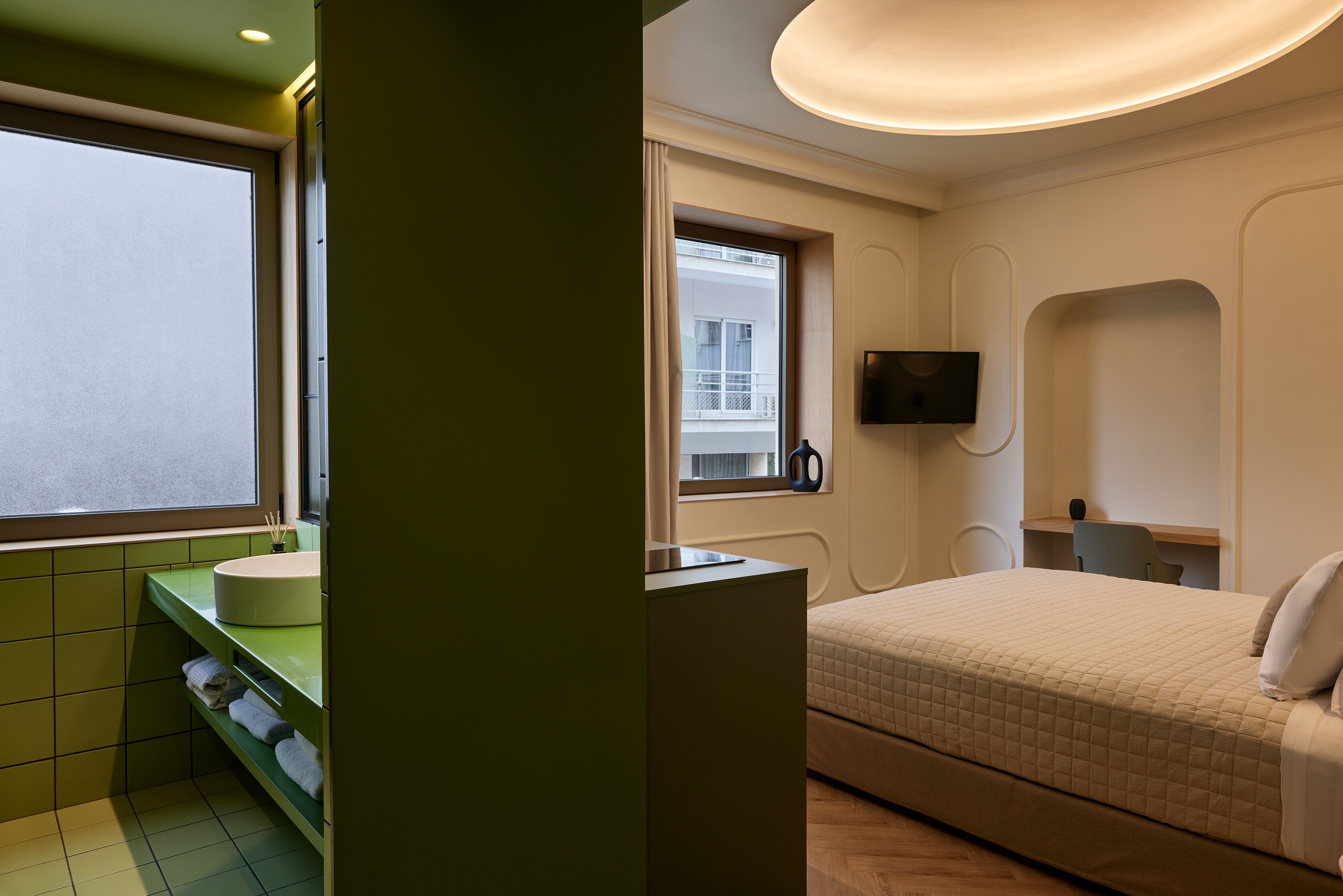 Une chambre d’hôtel moderne avec un grand lit, une coiffeuse verte, un téléviseur à écran plat et une fenêtre donnant sur l’immeuble.