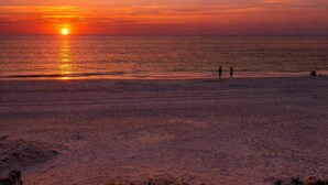 Escena de playa con un atardecer, dos personas caminando y una pequeña construcción en la arena.