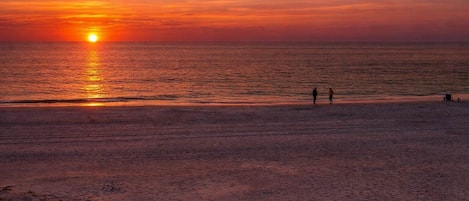 Escena de playa con un atardecer, dos personas caminando y una pequeña construcción en la arena.