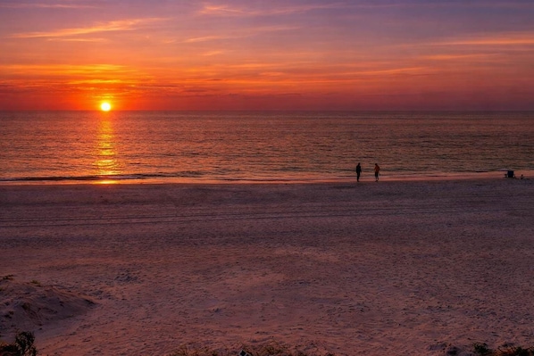 A beach scene with a sunset, two people walking, and a small structure on the sand.