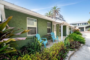 A row of green-framed buildings with a covered outdoor seating area, palm trees, and tropical plants.