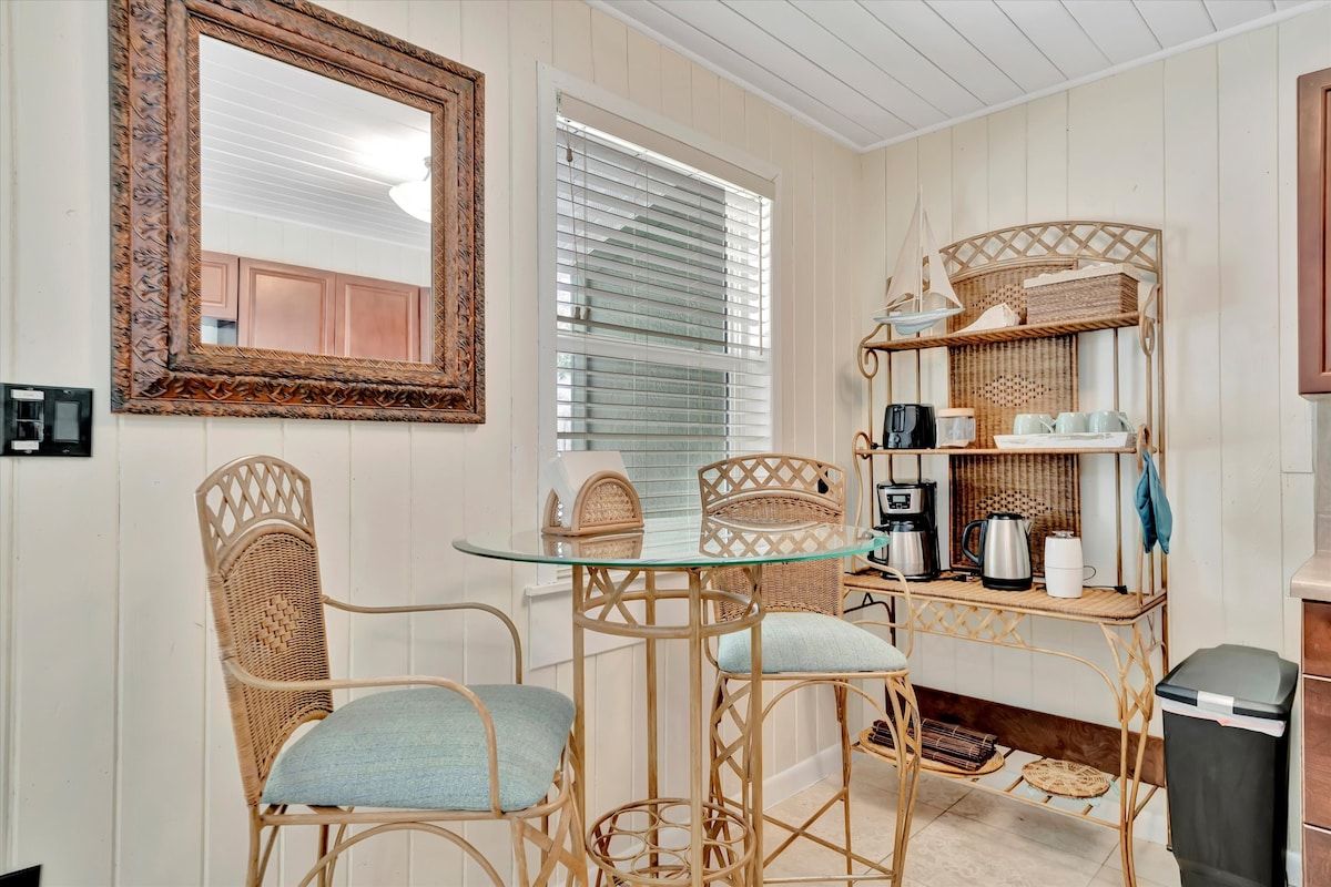 A compact kitchen with a glass table, wicker chairs, and a coffee station.