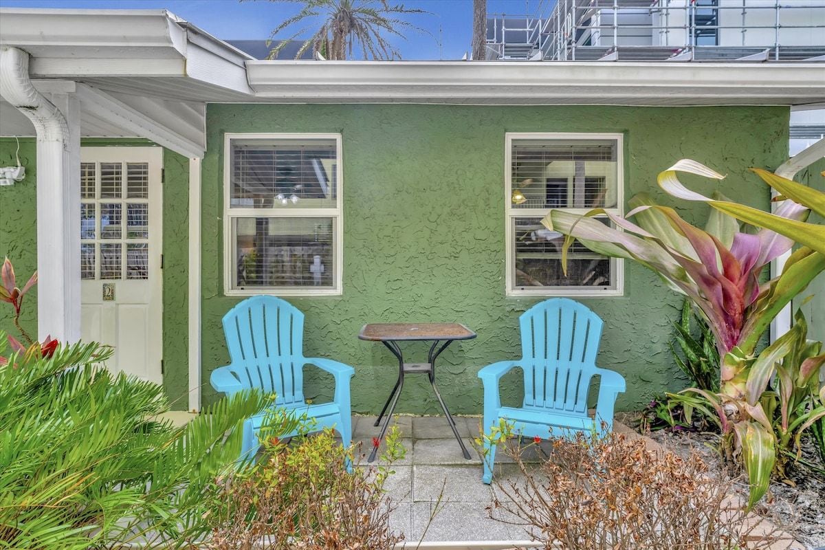 A charming green cottage with a welcoming front patio featuring turquoise Adirondack chairs and tropical plantings—your peaceful retreat awaits just steps from your front door.