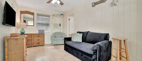 A room with a black sofa, a wicker side table, a wooden stool, a television, and a ceiling fan.