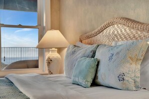A bed with a woven headboard, a lamp, and a window with a view of the sea. - 2B Condo Direct Beachfront Access & Ocean views! (Indian Rocks Beach)