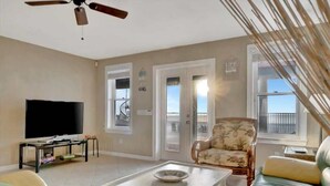 Un salon équipé d’une télévision, d’un ventilateur de plafond, d’un canapé, d’un fauteuil et d’une porte-fenêtre donnant sur un balcon.