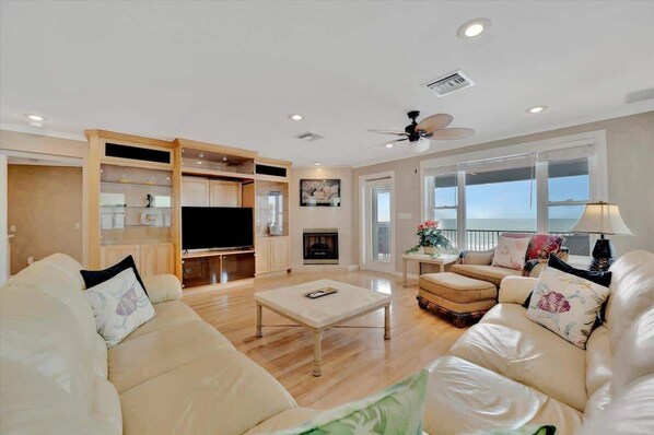 A spacious living room with a sofa, ottoman, and a coffee table. There is a fireplace, a television, and a ceiling fan.
