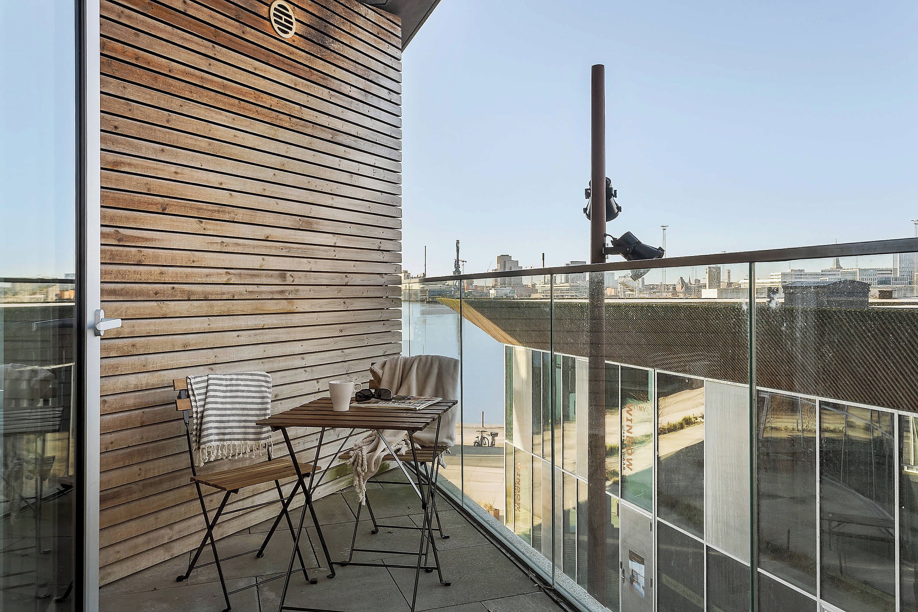 En balkon med trævæg, et bord og en stol, med udsigt over bybilledet.