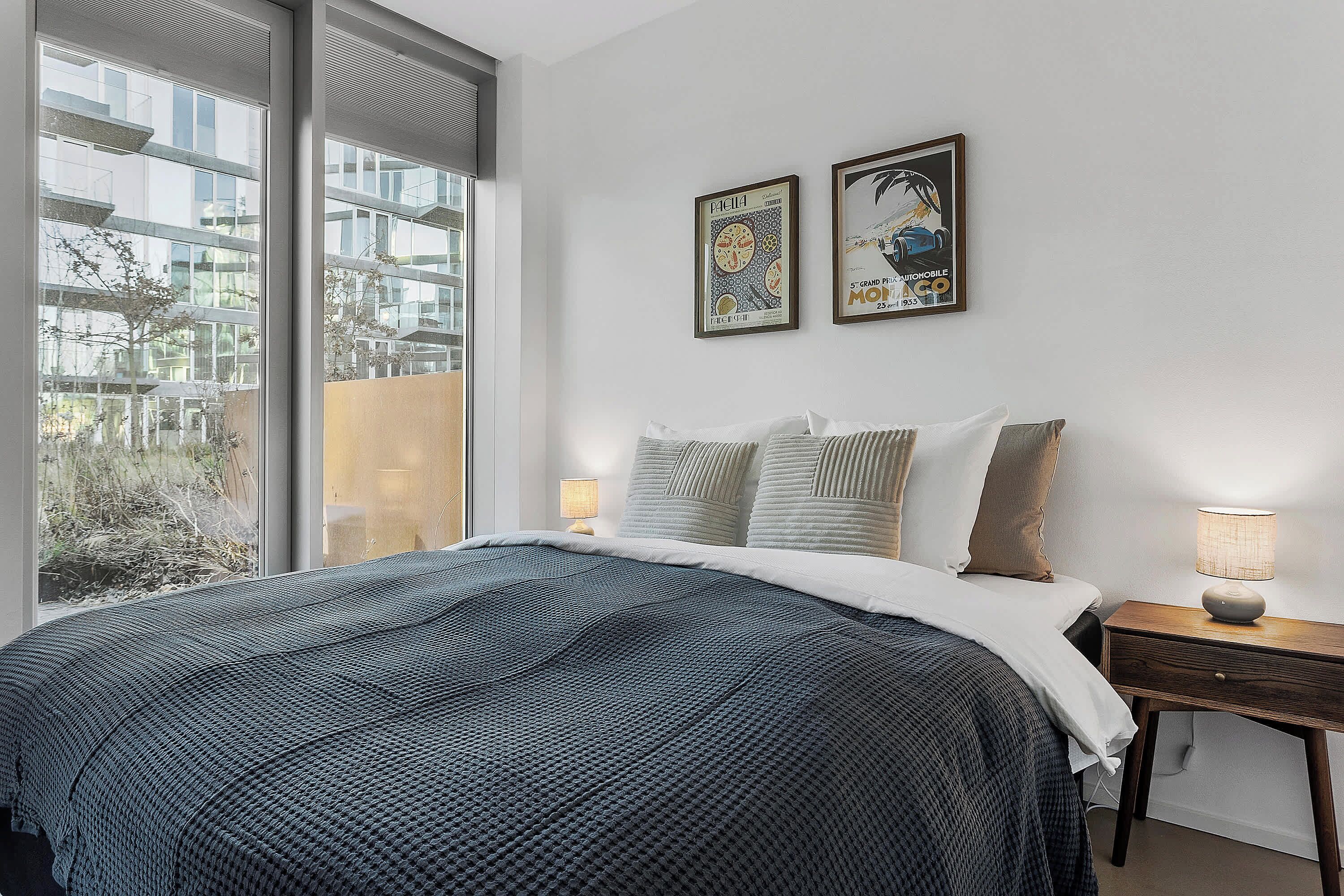 Un dormitorio con una cama grande, una mesita de noche con lámpara, dos cuadros en la pared y una ventana con vistas a edificios y árboles.