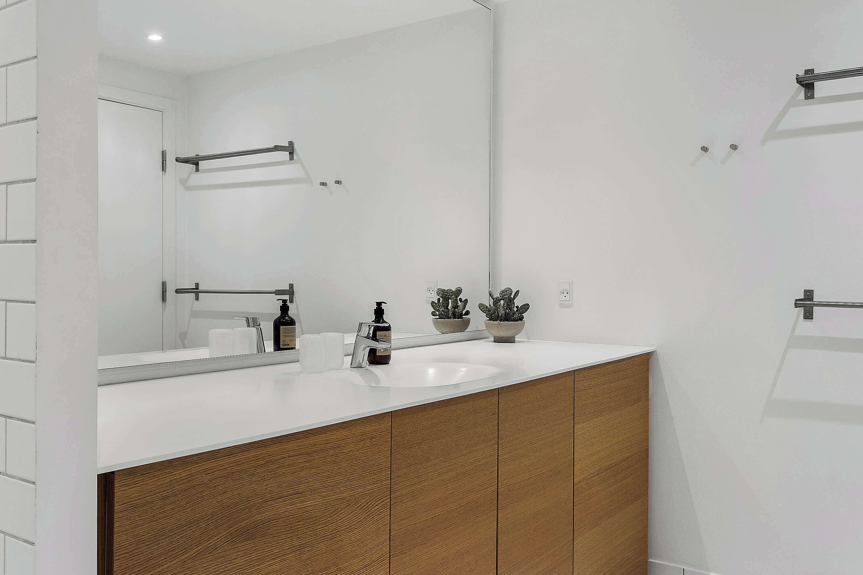 Un baño moderno con lavamanos blanco, armarios de madera, un espejo grande y dos plantas en macetas.