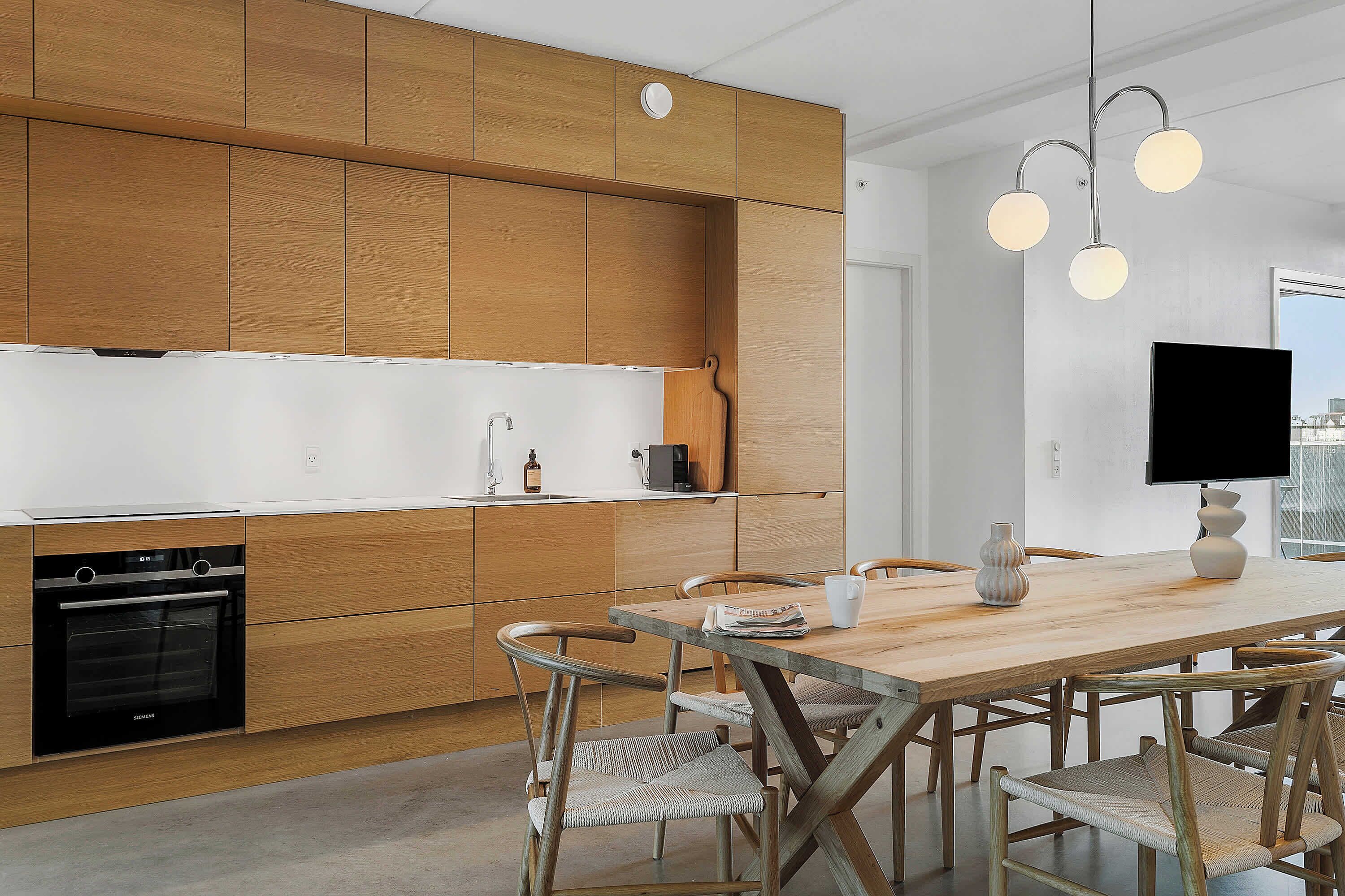 Eine moderne Küche mit Holzausstattung, einem eingebauten Backofen, einem Esstisch mit Stühlen und einem flachbildfernseher an der Wand.