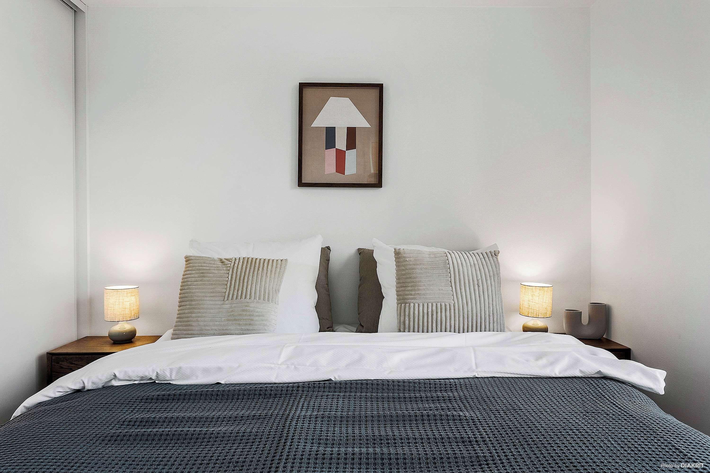A neatly made bed with white linens, two bedside lamps, and a framed abstract artwork on the wall.