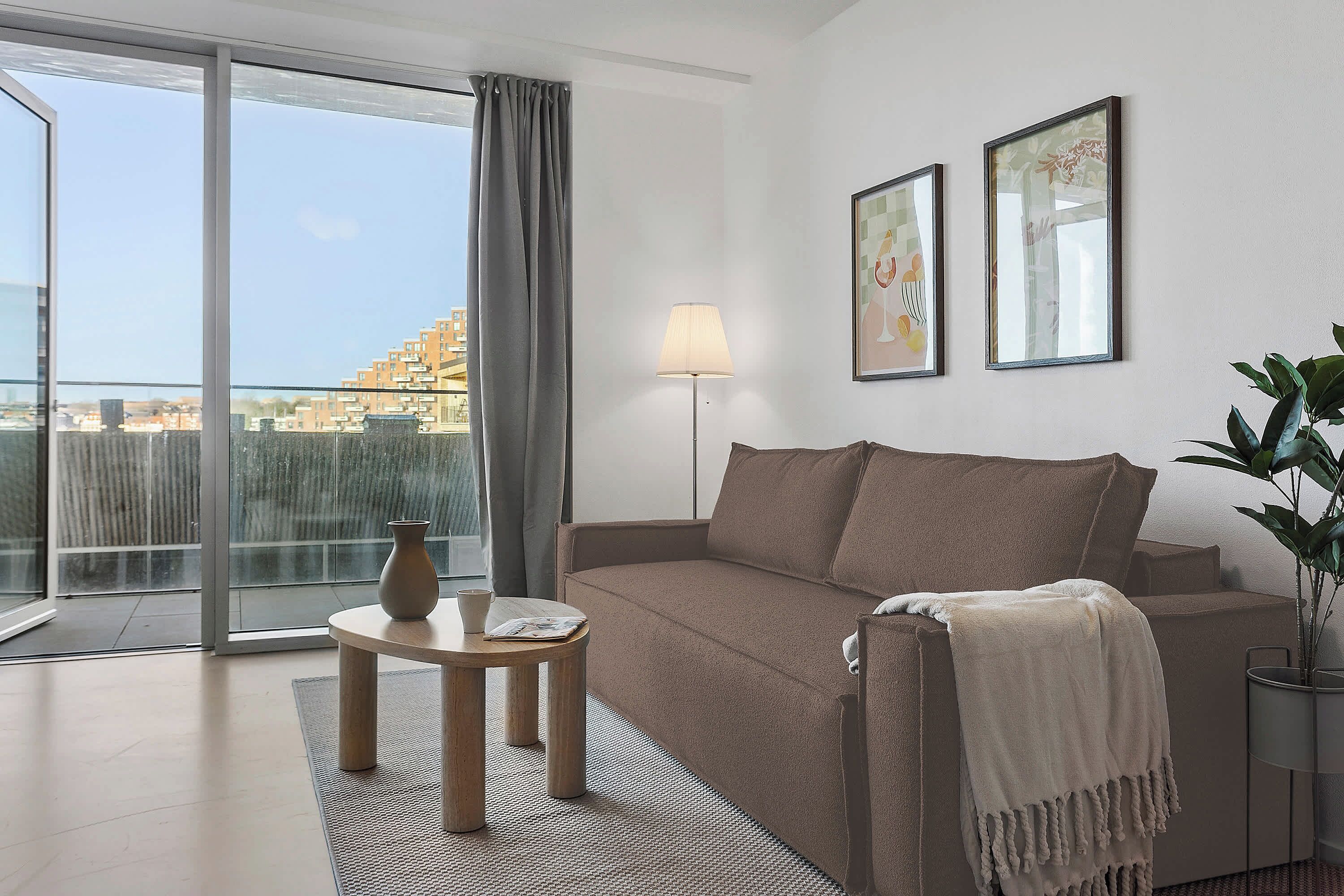 A modern living room with a sofa, a table, a lamp, and a view through a large window.