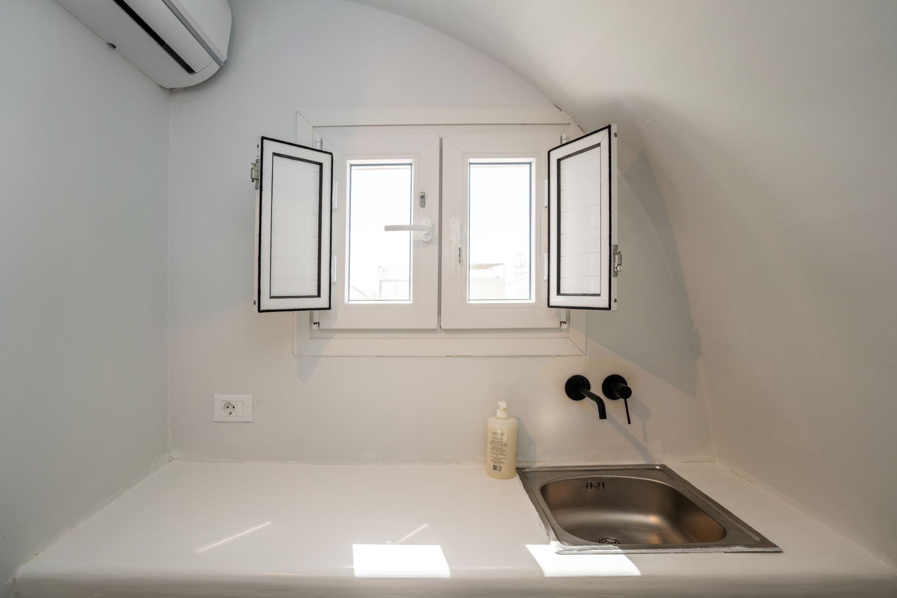 Une petite cuisine blanche, avec un seul évier en acier inoxydable, une fenêtre dont les volets sont ouverts, et un flacon de savon liquide posé sur le comptoir.