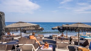 A beach with lounge chairs and umbrellas, a clear sky, and the ocean in the background. - Zen Suites & Spa (Santorini)