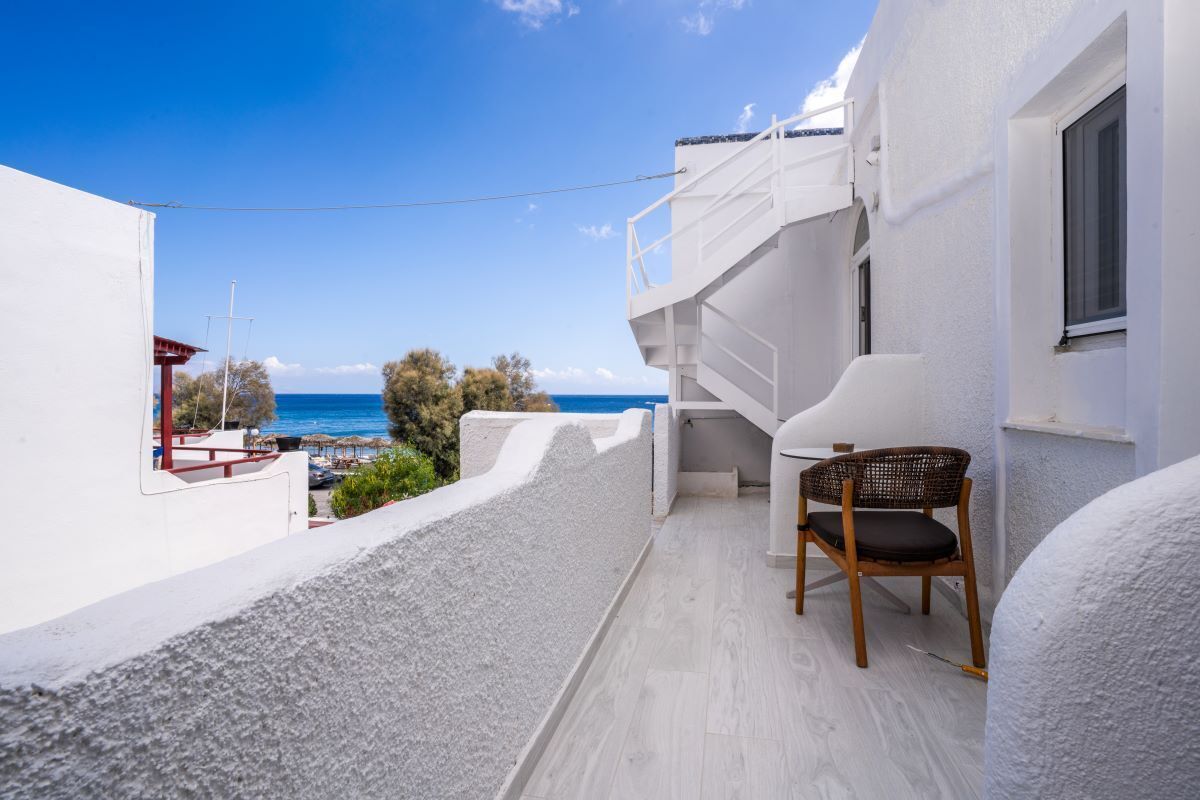 A narrow balcony with a view of the sea, a white building, and a wooden chair.