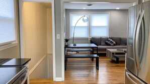 A modern kitchen with a stainless steel refrigerator, wooden flooring, a black dining table, and a grey sofa in the background.