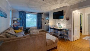 A living room with a sofa, a coffee table, a TV mounted on the wall, and a window with curtains.