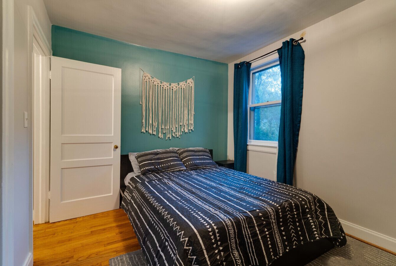 A bedroom with a bed, a window with curtains, a door, and a wall with a macrame decoration.