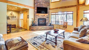 A spacious living room with a stone fireplace, leather furniture, and large windows offering views of the outdoors. - Fire Pit: Cozy Winter Cabin, 45 Mi to Sunrise Park (Show Low)
