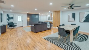 A modern living room with a sofa, dining table, and chairs. There is a wooden floor and a ceiling fan.