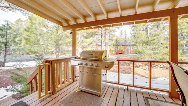 A covered outdoor deck with a stainless steel grill, wooden railings, and a view of a forested area.