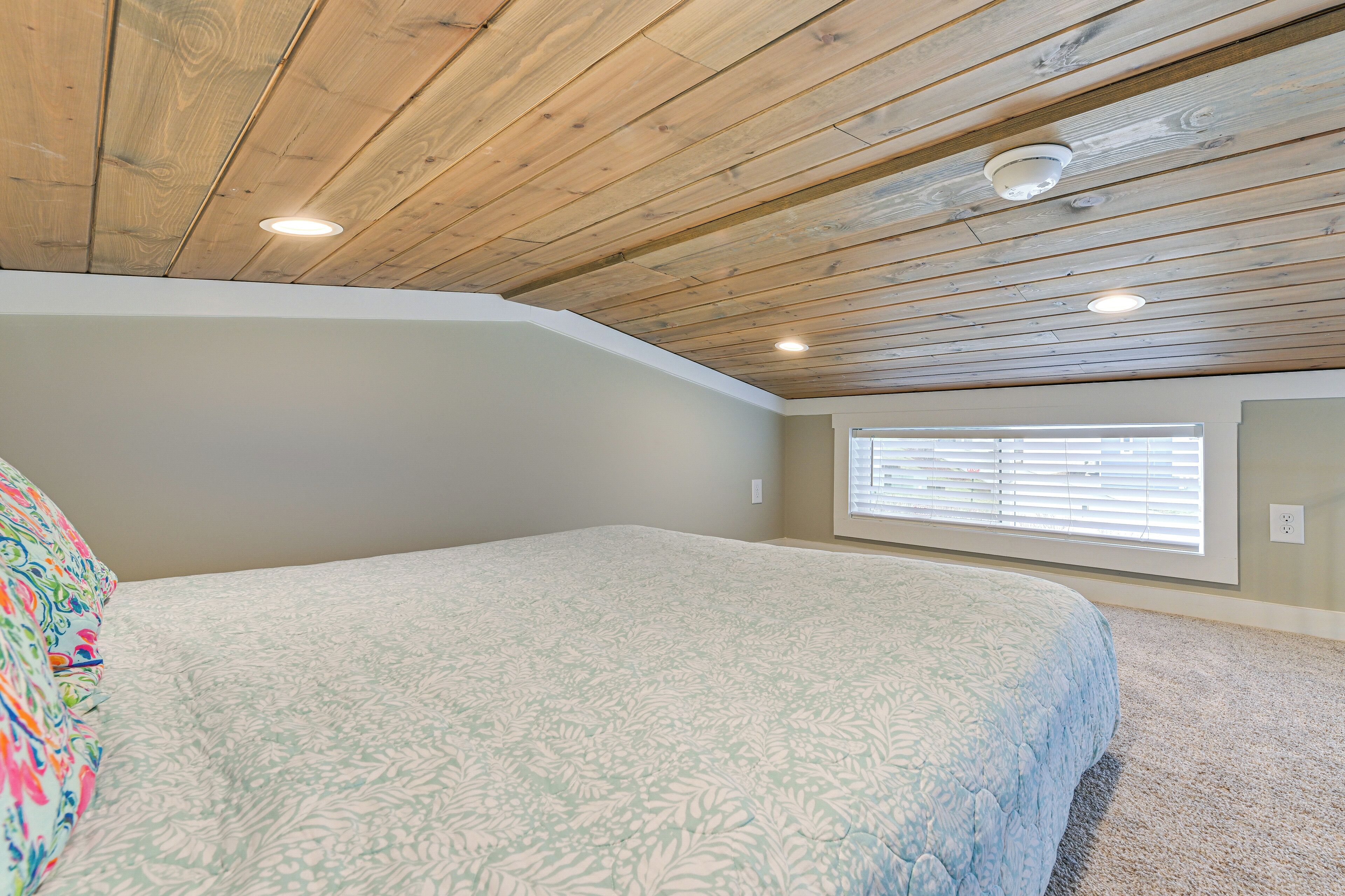 Une chambre avec un plafond en bois, une fenêtre avec des stores et un lit recouvert d’une couette à motifs floraux.