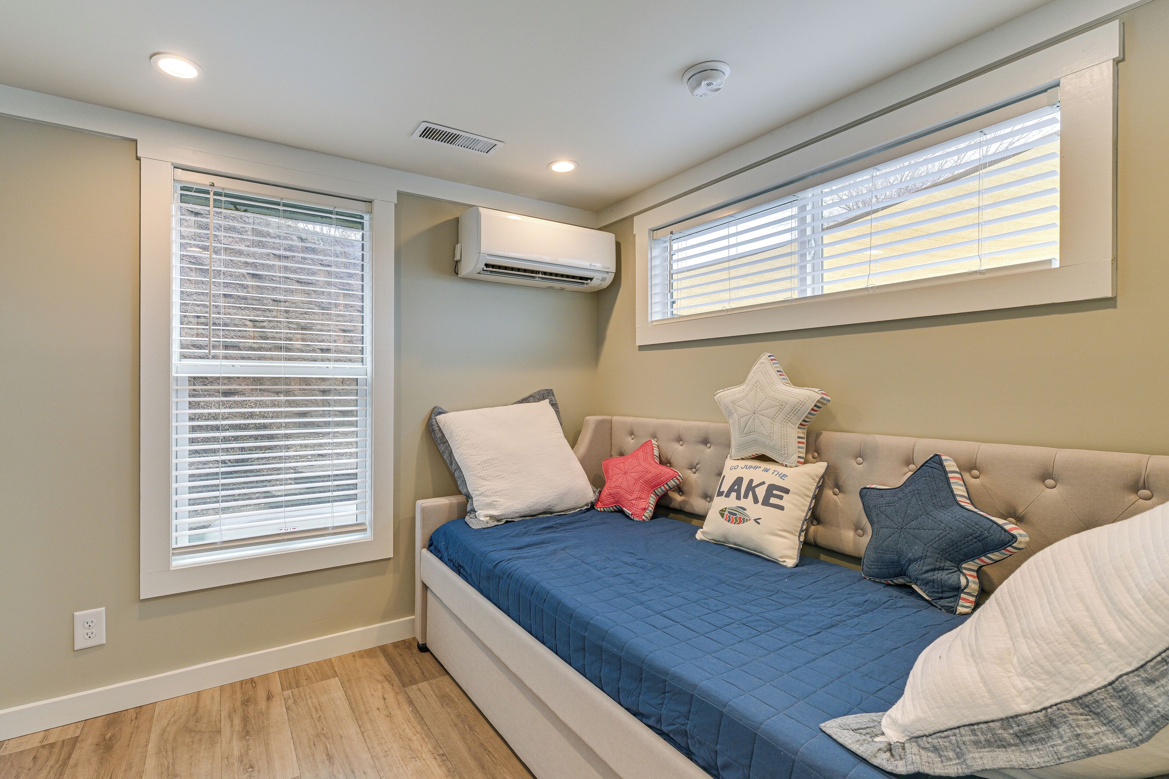 A bedroom with a bed, two windows with blinds, an air conditioning unit, and a ceiling light.