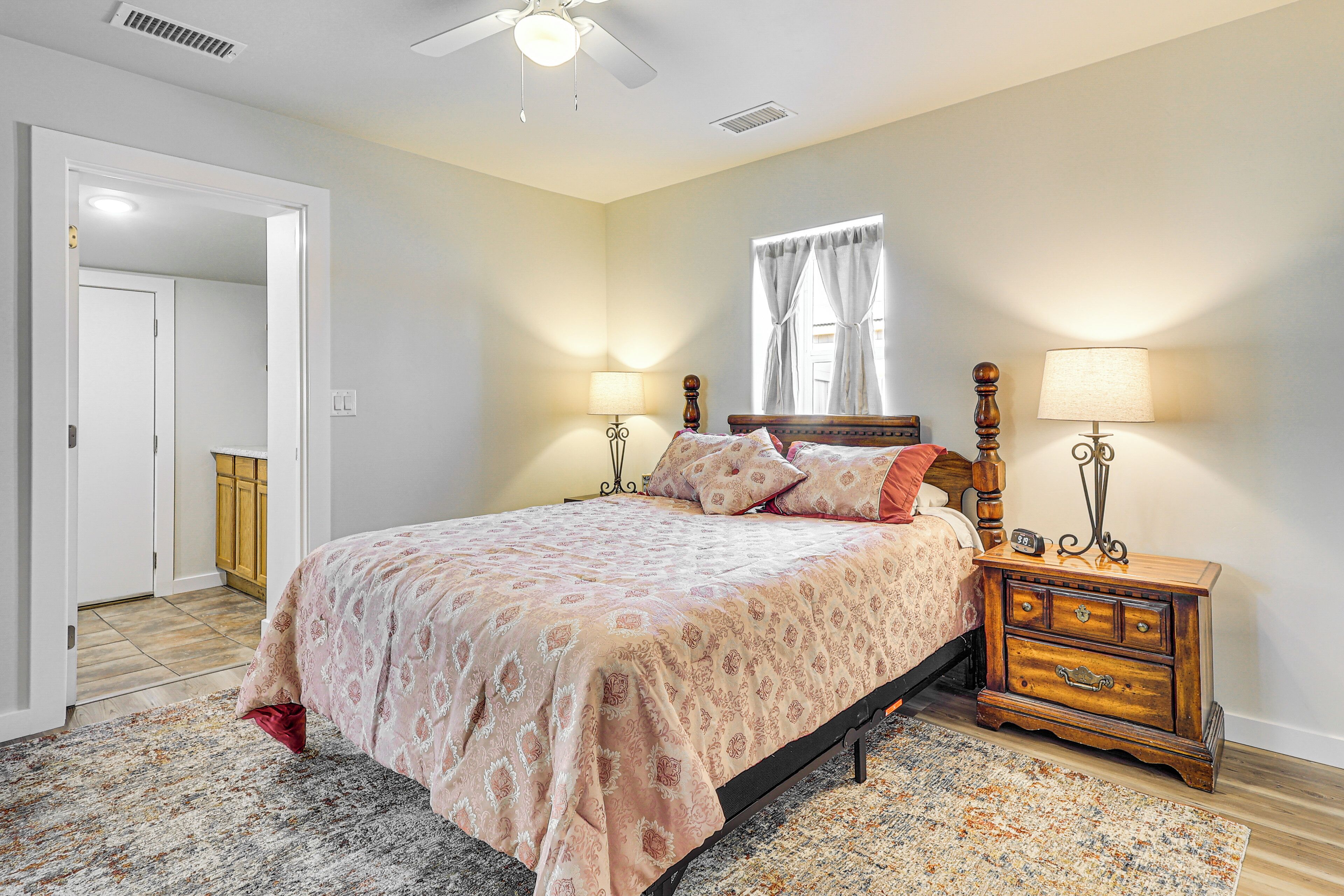 A bedroom with a bed, bedside tables, a nightstand, a window with curtains, and a door leading to another room.