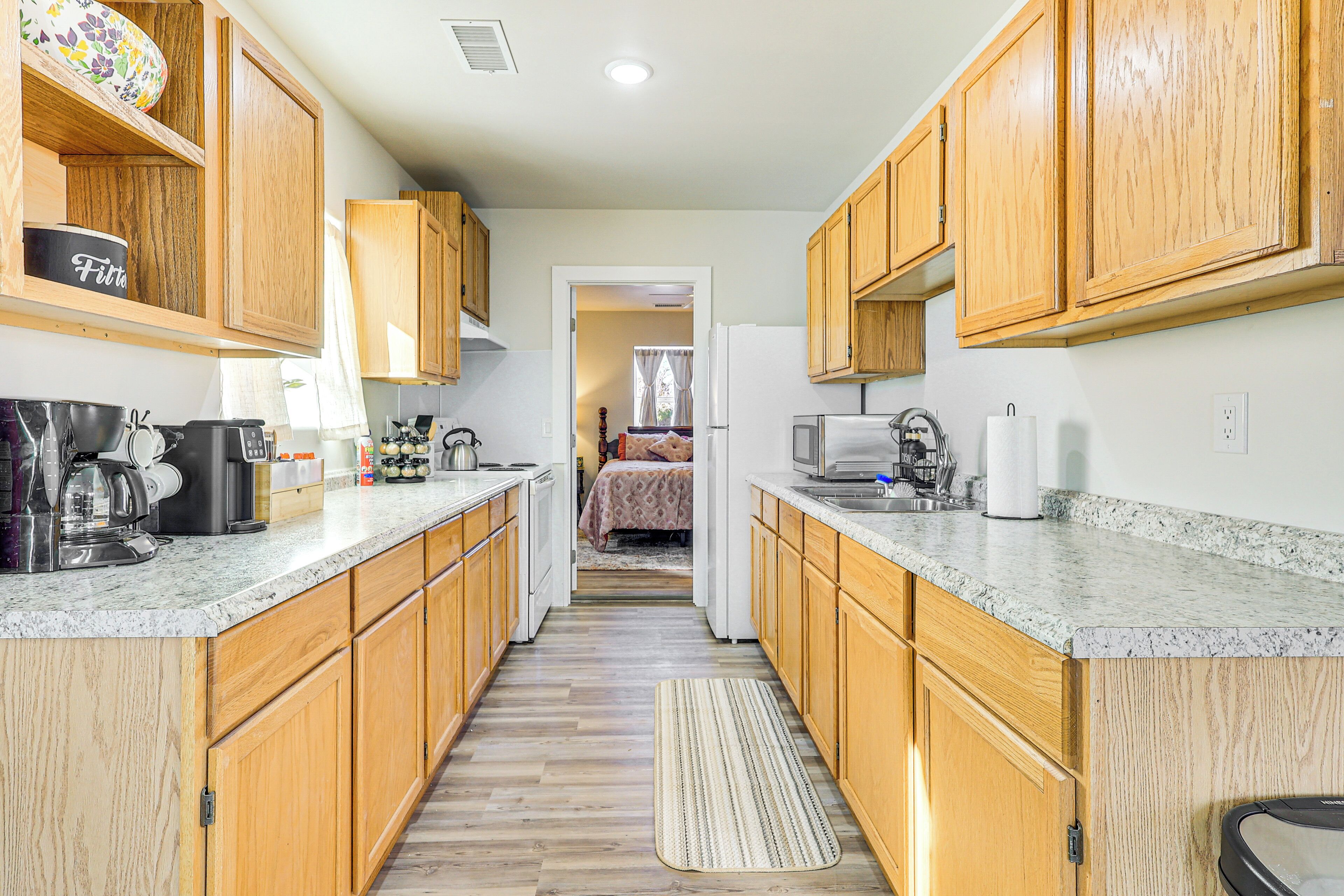 A narrow kitchen with wooden cabinets, granite countertops, and a view into a bedroom.