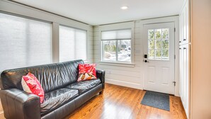 Una habitación con un sofá de cuero negro, dos almohadas rojas, suelo de madera, una puerta blanca y una ventana con persianas.