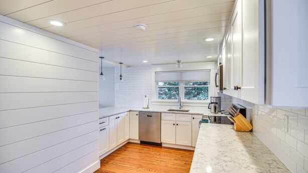 Una cocina moderna con armarios blancos, lavavajillas, pileta y ventana con vista a árboles.