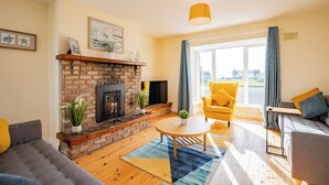 Ein Wohnzimmer mit Kamin aus Backstein, einem gelben Sessel, einem grauen Sofa und einem großen Fenster mit Vorhängen.